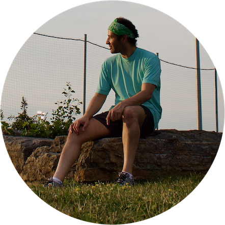 Rodrigo sitting on a rock in a park staring at the horizon at sunset. Rodrigo is wearing shorts, t-shirt, running shoes and a green bandana, looking as if resting after doing some exercise in the afternoon.