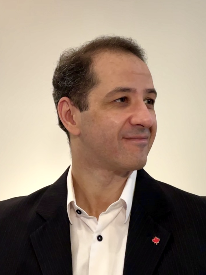 Rodrigo sitting on a chair, wearing a dark jacket and a white shirt without tie, looking to his left and smiling. On his lapel there is a pin in the shape of a red maple leaf.